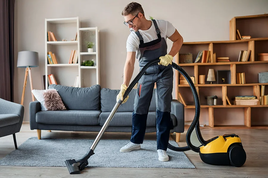 young man smiling, wearing glasses, wearing a pinny, vacuuming the living room carpet