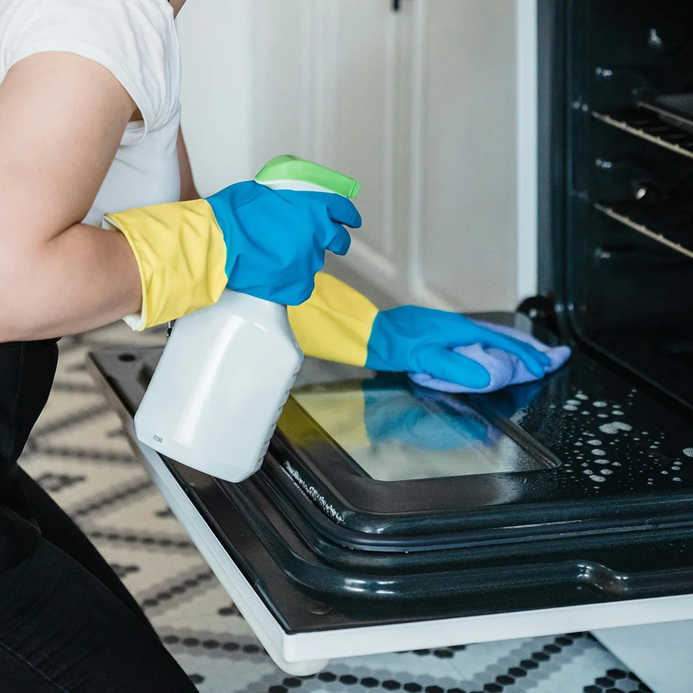 A picture of a cleaner wearing rubber gloves holding cleaning spray and wiping clean an oven.